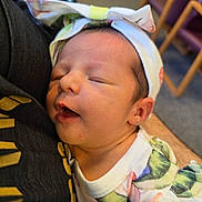 Eliana is registered to the contest to win money with this photo: baby, background, bow, candid, caregiver, chairs, close_up, colorful_clothing, face, family, headband, held, indoors, infant, newborn, onesie, peaceful, portrait, sleeping, warm_lighting