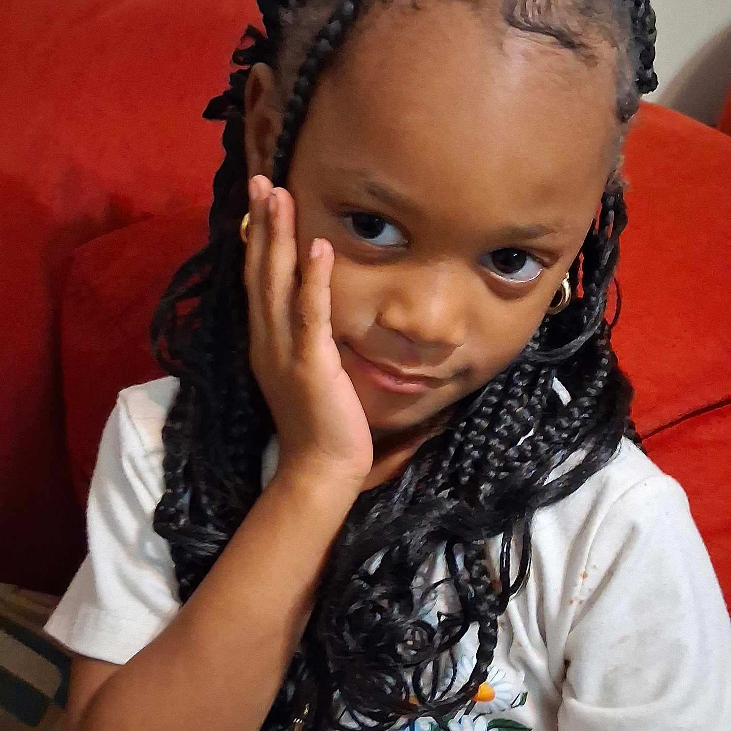 Kadaisa is registered to the contest to win money with this photo: braided_hair, casual_clothing, child, cozy, cute, daisy_print, earrings, expression, girl, hand_on_face, home, indoor, person, portrait, red_couch, relaxed, seated, smile, white_shirt, young