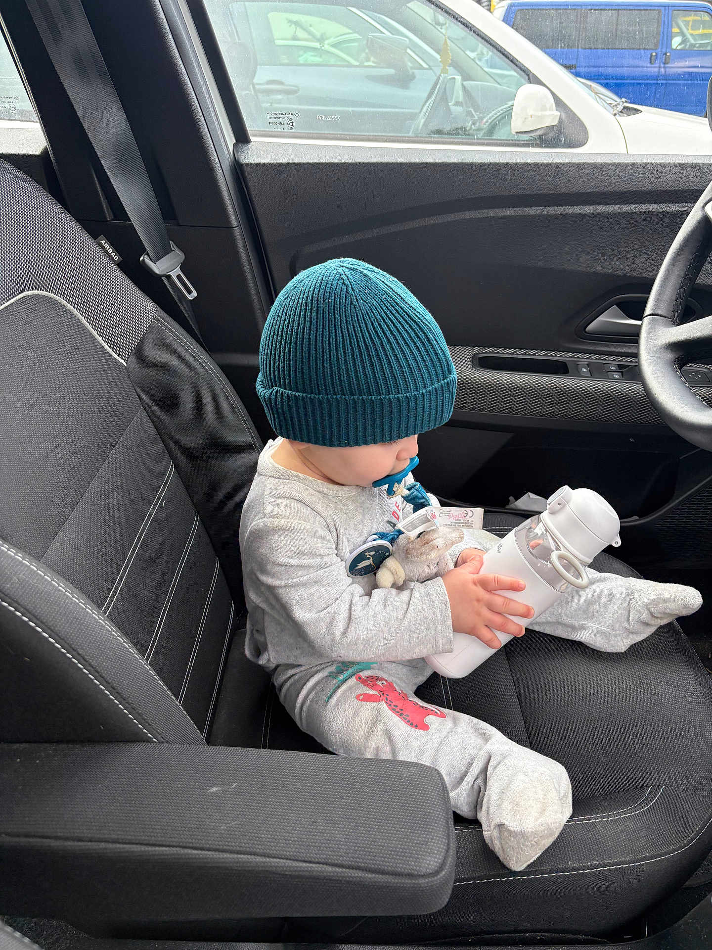 Liam participe au concours pour gagner de l'argent avec cette photo : child, infant, baby, beanie, hat, pacifier, water_bottle, plush_toy, car_interior, car_seat, seatbelt, armrest, steering_wheel, window, gray_clothing, sitting, passenger, toddler, cozy_outfit, front_seat