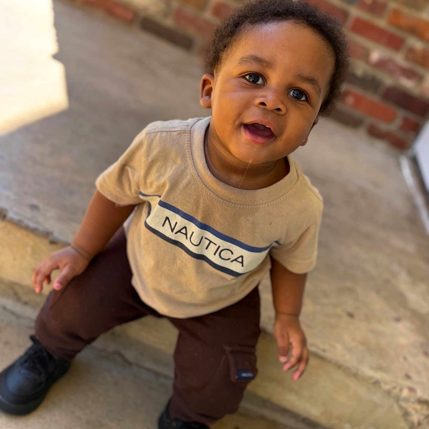 Zamir is registered to the contest to win money with this photo: toddler, child, boy, smiling, curious, outdoor, porch, brick_wall, clothing, shirt, pants, black_shoes, face, eyes, skin, hair, concrete, daylight, casual, portrait