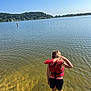 back_view, casual_clothing, child, daytime, lake, life_jacket, nature, outdoor, person, recreation, reflection, scenic, shoreline, sky, summer, sunlight, trees, wading, water, water_surface