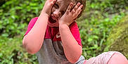 Leah is registered to the contest to win money with this photo: child, forest, greenery, moss, nature, sitting, playful, hands_on_face, expression, casual_clothing, cropped_pants, white_shoes, outdoor, daylight, young, person, vegetation, leaves, tree, fun