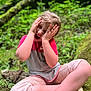 child, forest, greenery, moss, nature, sitting, playful, hands_on_face, expression, casual_clothing, cropped_pants, white_shoes, outdoor, daylight, young, person, vegetation, leaves, tree, fun