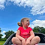 child, girl, sitting, cross_legged, red_shirt, colorful_socks, outdoor, blue_sky, clouds, trees, greenery, vehicle, casual, daylight, nature, young_child, curly_hair, playful, relaxed, portrait
