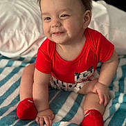 Adriel Romero joined the competition — help win amazing prizes! baby, child, smiling, red_shirt, red_socks, striped_blanket, indoor, cute, happy, sitting, person, infant, cozy, soft_lighting, portrait, hand, footwear, clothing, bed, young