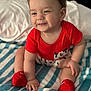 baby, child, smiling, red_shirt, red_socks, striped_blanket, indoor, cute, happy, sitting, person, infant, cozy, soft_lighting, portrait, hand, footwear, clothing, bed, young