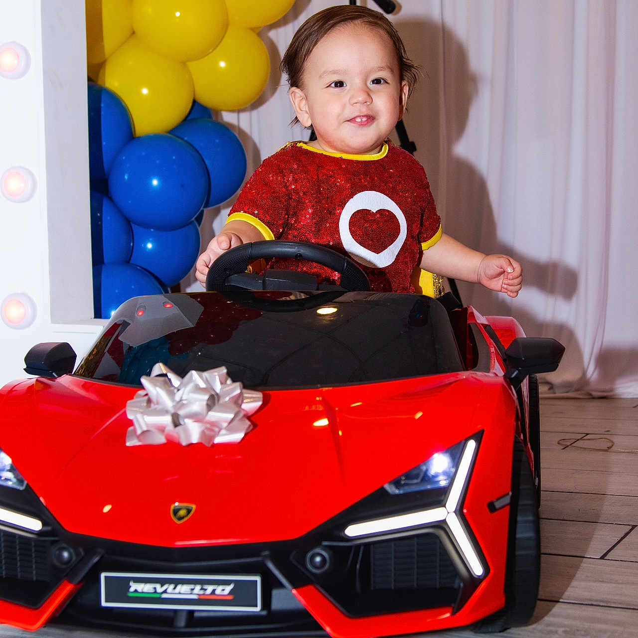 Adriel Romero joined the competition — help win amazing prizes! balloons, blue_balloons, child, curtains, decor, face, flooring, gift_bow, hands, happy, indoor, person, photography, red_car, smiling, sparkly_clothing, toddler, toy_car, tripod, yellow_balloons