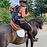 woman, person, horse, dog, rider, equestrian, backpack, goggles, helmet, sunglasses, outdoor, trees, flowers, street, saddle, bridle, reins, riding_boots, animal, portrait