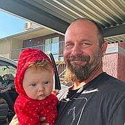 Lilah joined the competition — help win amazing prizes! baby, man, beard, red_hoodie, polka_dots, outdoor, car, building, metal_canopy, smile, portrait, holding, child, casual_clothing, daylight, facial_expression, person, headwear, window, door