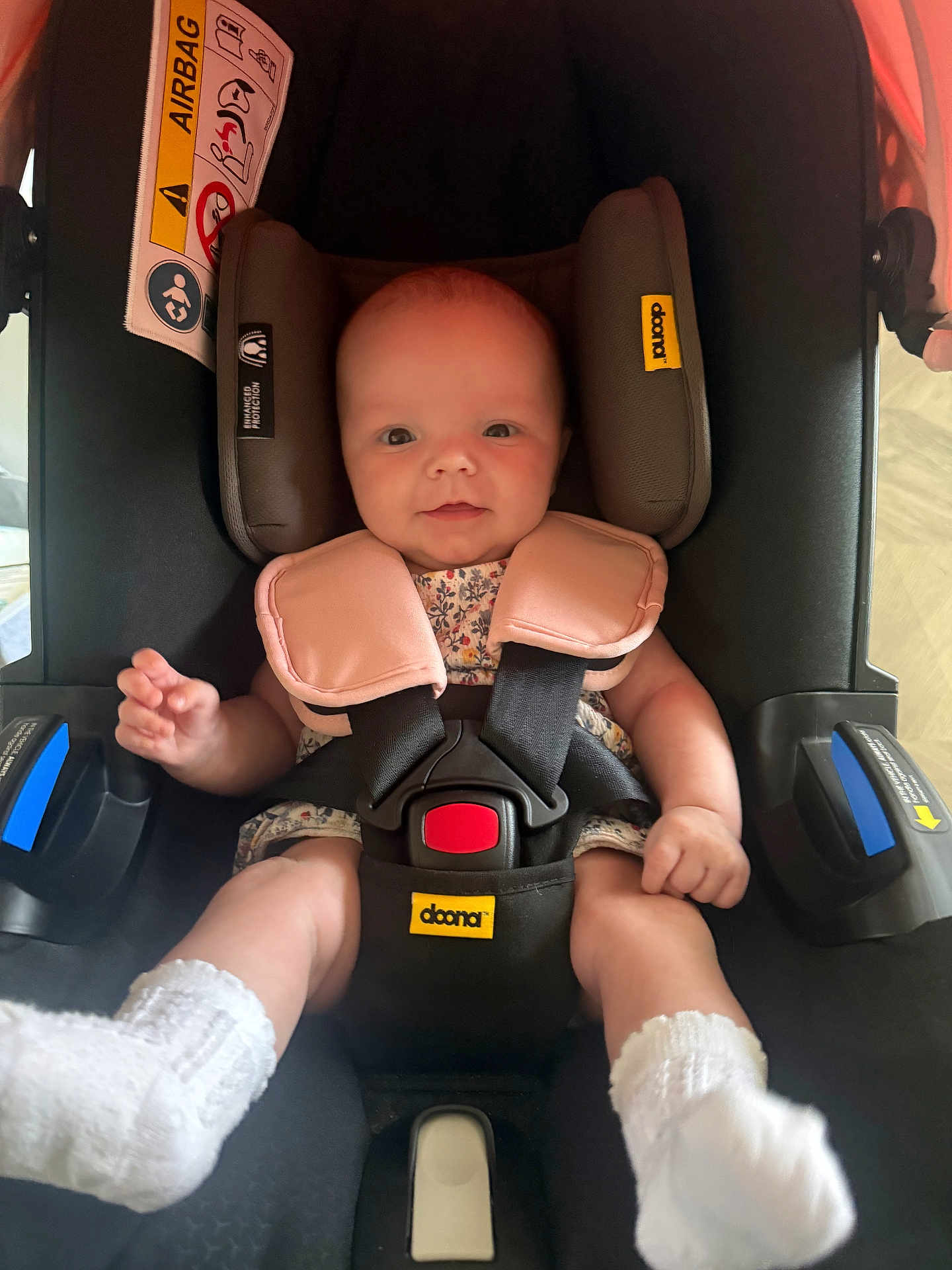 Bonnie is registered to the contest to win money with this photo: baby, car_seat, infant, safety_harness, white_socks, floral_outfit, red_buckle, black_car_seat, yellow_label, child, cute, smiling, infant_face, seatbelt, indoors, person, baby_clothing, head_support, small_child, baby_feet