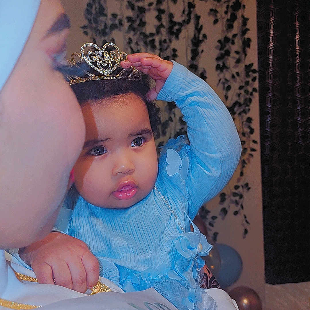Mariam is registered to the contest to win money with this photo: baby, balloons, blue_clothing, celebration, child, closeup, curious, decor, event, face, family, hand, headwear, holding, indoor, person, portrait, tiara, toddler, woman