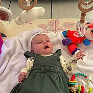 Giulia participe au concours pour gagner de l'argent avec cette photo : baby, infant, child, blanket, toy, dress, headband, flower, green_dress, white_tights, soft_toy, colorful_toys, play_area, floor_tiles, cute, smiling, indoors, person, lying_down, happy