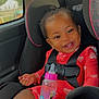 baby_bottle, car_seat, child, daylight, dimple, face, hair, hand, happy, pattern, pink, red_clothing, safety, seatbelt, sitting, smile, toddler, toy, vehicle_interior, window