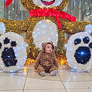 Jawad is registered to the contest to win money with this photo: baby, brown, child, costume, curious, cute, decoration, face, festive, floor, holiday, indoor, lights, plush, red_scarf, sitting, smile, teddy_bear, toy, white_lights