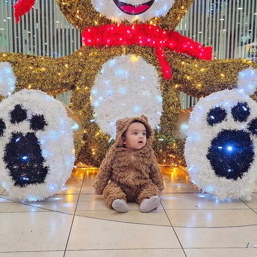 Jawad is registered to the contest to win money with this photo: baby, brown, child, costume, curious, cute, decoration, face, festive, floor, holiday, indoor, lights, plush, red_scarf, sitting, smile, teddy_bear, toy, white_lights