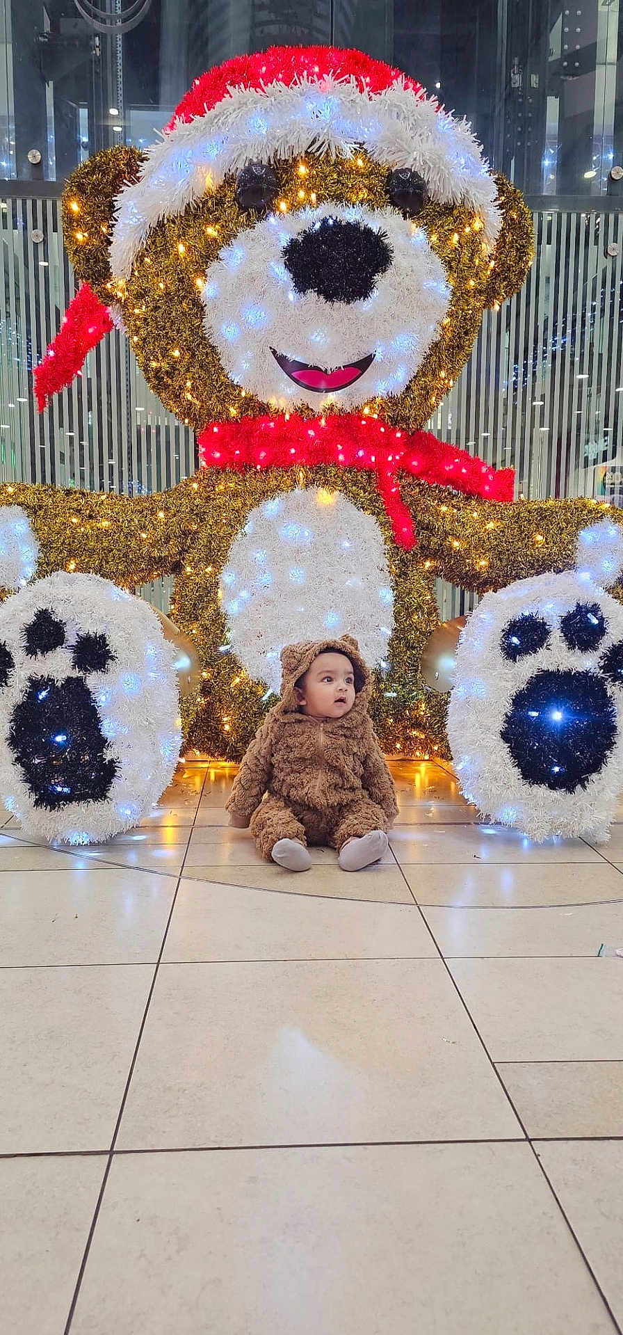 Jawad is registered to the contest to win money with this photo: baby, brown, child, costume, curious, cute, decoration, face, festive, floor, holiday, indoor, lights, plush, red_scarf, sitting, smile, teddy_bear, toy, white_lights
