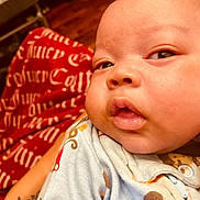 Zakari is registered to the contest to win money with this photo: baby, close_up, face, infant, child, onesie, patterned_clothing, cheeks, mouth, eyes, skin, red_blanket, text_on_blanket, tattoo, hand, wooden_floor, indoors, person, portrait, curious