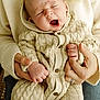 Archie is registered to the contest to win money with this photo: baby, yawning, knitted_outfit, cream_color, hands, adult, holding, cozy, warm, cuddly, portrait, indoor, closeup, soft_lighting, human, infant, sitting, sweater, buttons, skin