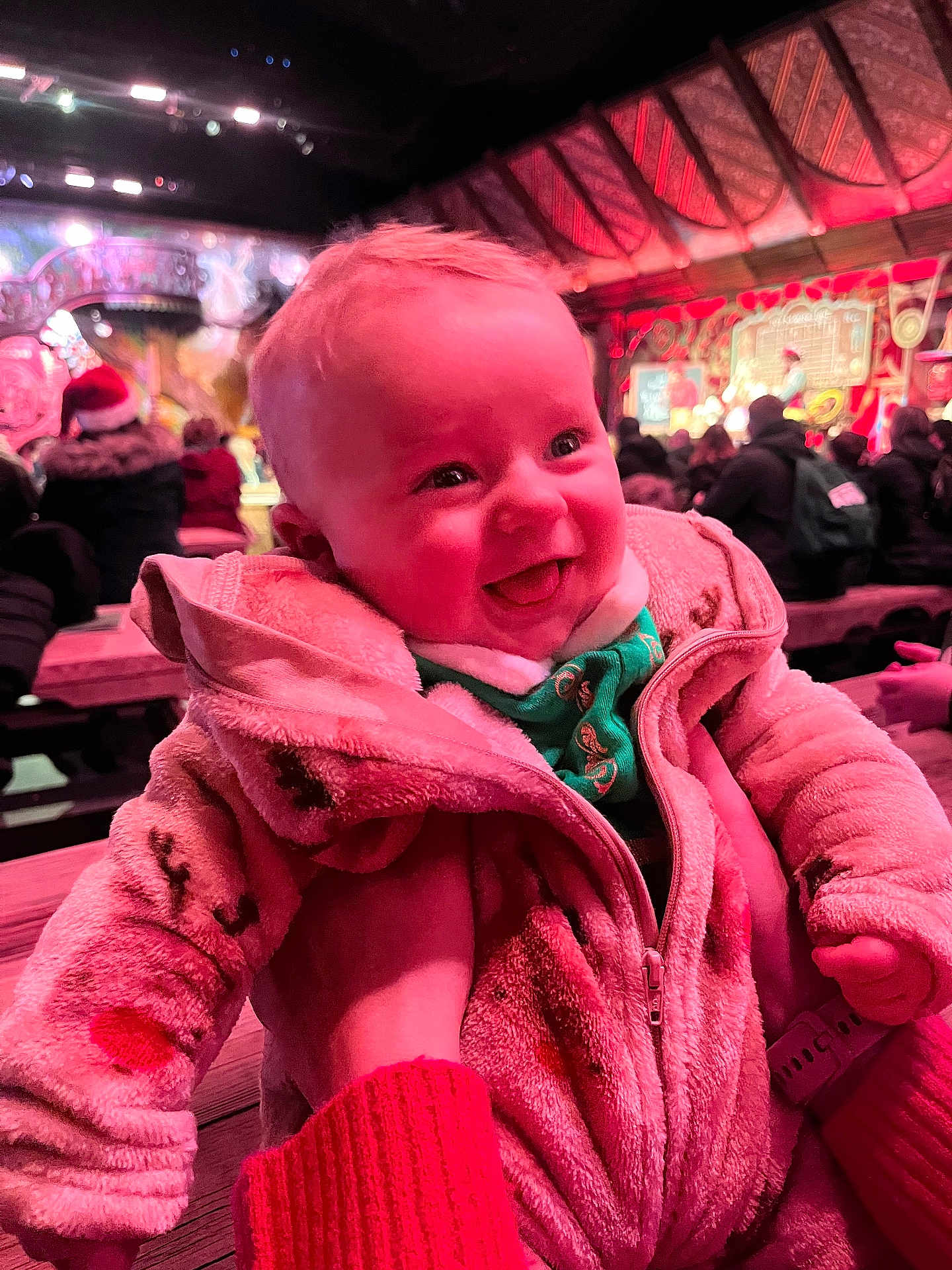 Archie is registered to the contest to win money with this photo: baby, child, smiling, jacket, pink, happy, indoor, festive, people, crowd, warm_clothing, portrait, infant, cute, holding, bench, event, colorful_lighting, face, person