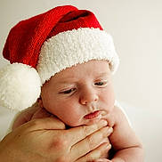 Archie is registered to the contest to win money with this photo: baby, santa_hat, red_hat, white_fur, face, hand, skin, closeup, expression, cute, child, infant, portrait, holiday, christmas, soft_light, neutral_background, holding, pensive, young