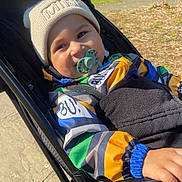 Elijah is registered to the contest to win money with this photo: toddler, child, stroller, pacifier, beanie, jacket, outdoor, park, sunlight, smile, hand, colorful_clothing, seatbelt, nature, trees, pathway, baby, face, person, daylight