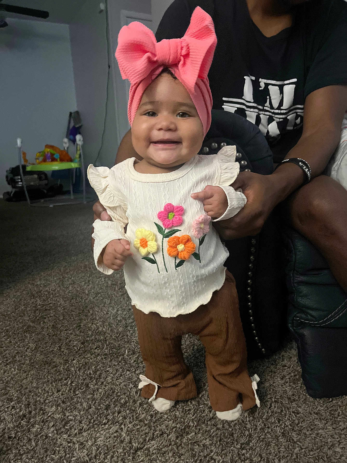 Fayth joined the competition — help win amazing prizes! baby, child, smiling, pink_bow, headband, white_sweater, flower_embroidery, brown_pants, carpet, indoor, person, adult_hands, happy, standing, home, furniture, black_tshirt, toy, cozy, cute