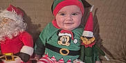 Owen participe au concours pour gagner de l'argent avec cette photo : baby, child, smile, festive_clothing, elf_hat, striped_pants, plush_toy, santa_claus_doll, elf_doll, couch, indoor, holiday, christmas, happy, cute, person, seated, cozy, red, green