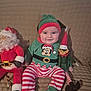 baby, child, smile, festive_clothing, elf_hat, striped_pants, plush_toy, santa_claus_doll, elf_doll, couch, indoor, holiday, christmas, happy, cute, person, seated, cozy, red, green