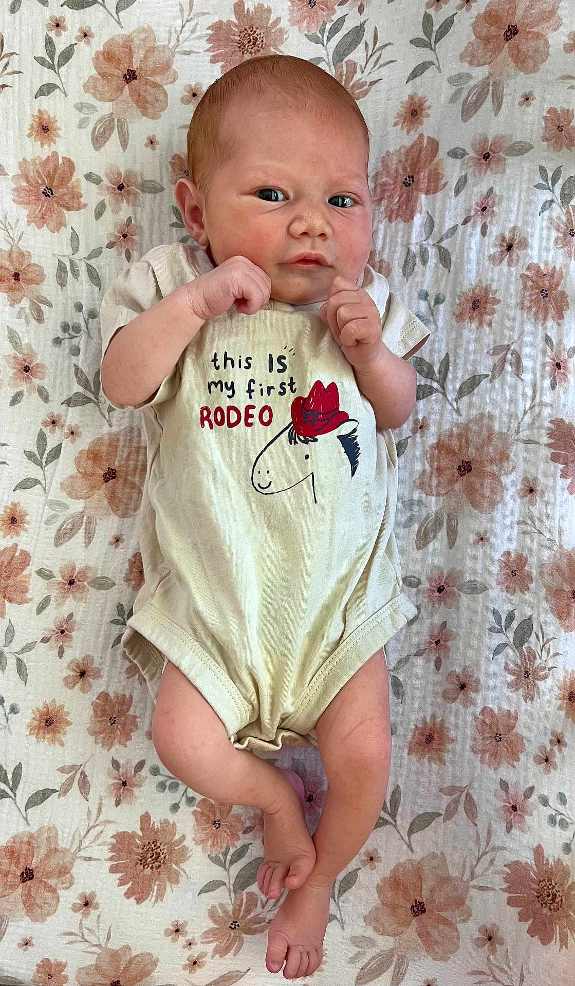 Genevieve is registered to the contest to win money with this photo: baby, newborn, infant, onesie, clothing, floral_pattern, blanket, lying_down, cute, face, hands, feet, red_hat_cartoon, text, skin, person, indoors, soft_lighting, resting, expression