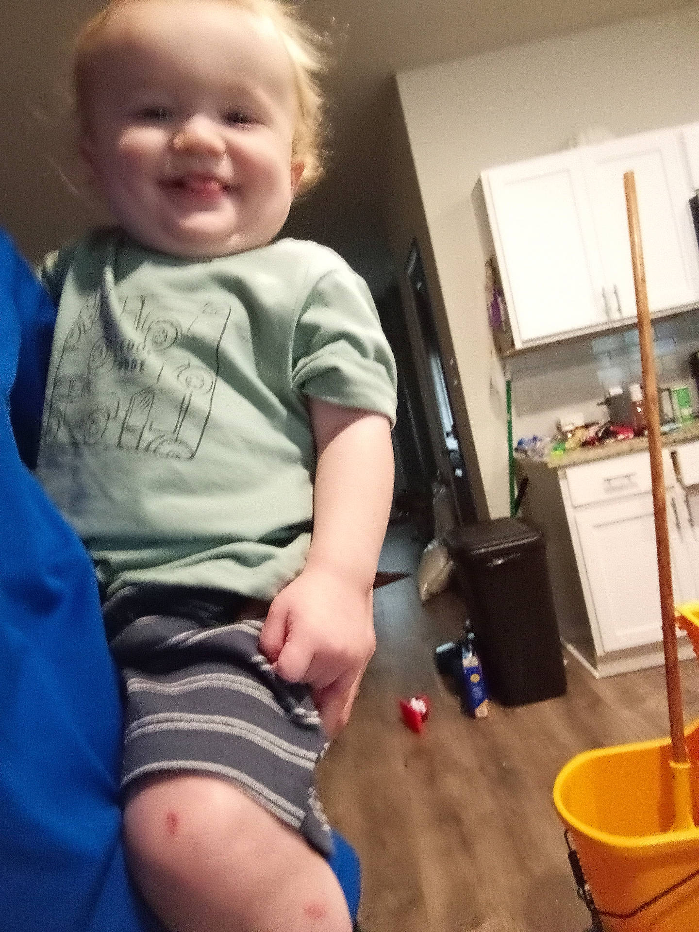 blurry, cabinet, casual, child, face, floor, hand, home, indoor, kitchen, light, mop_bucket, person, shorts, smile, toddler, toy, trash_can, tshirt, wood_floor