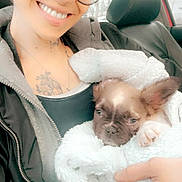 Frankie James is registered to the contest to win money with this photo: person, smiling, glasses, beanie, puppy, dog, blanket, car, seat, hand, jacket, tattoo, cozy, cute, pet, indoor, portrait, happy, young_adult, casual