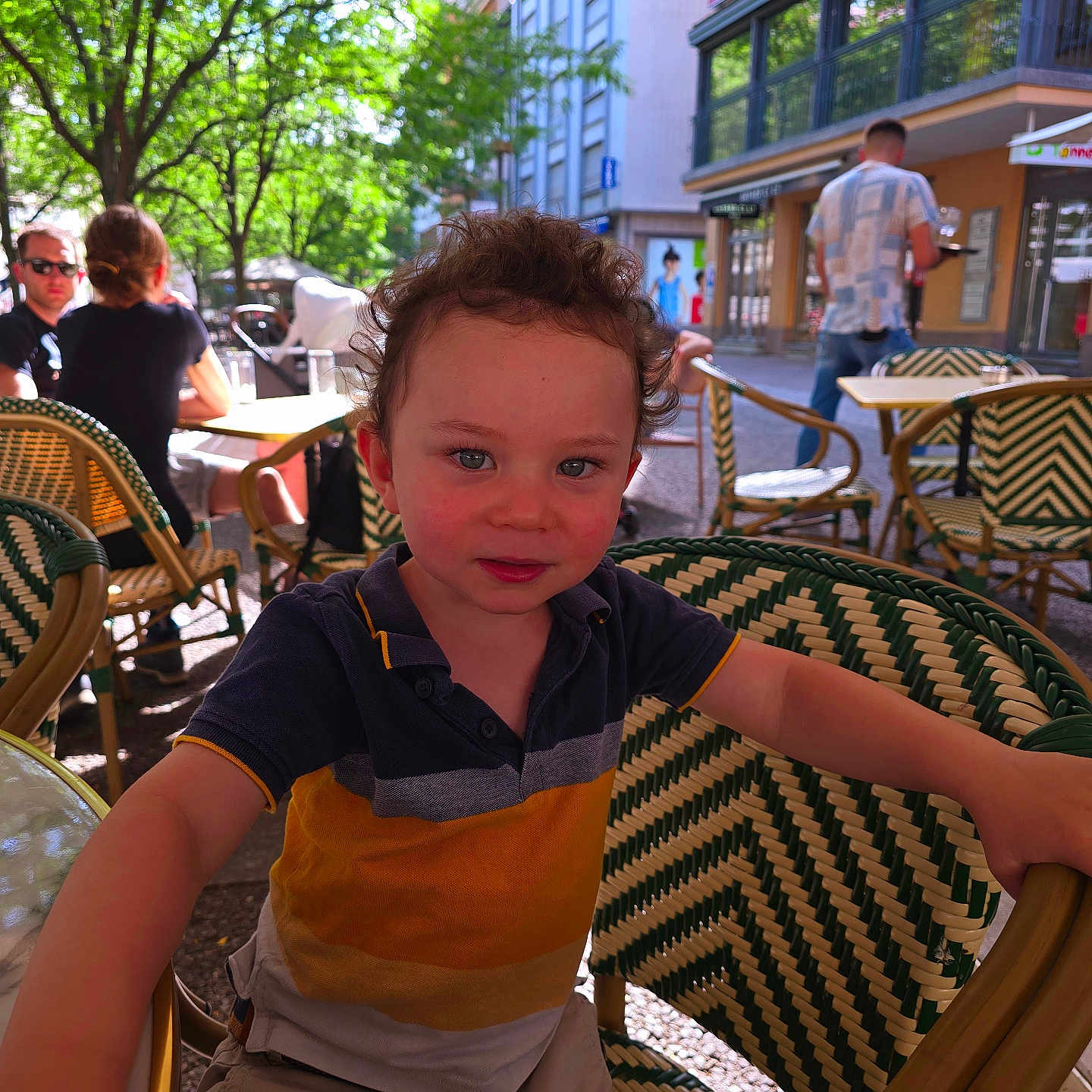 Vicente participe au concours pour gagner de l'argent avec cette photo : building, cafe, casual_clothing, chair, child, curious, curly_hair, daytime, outdoor, people, person, relaxing, restaurant, smiling, street, sunlight, table, toddler, trees, urban