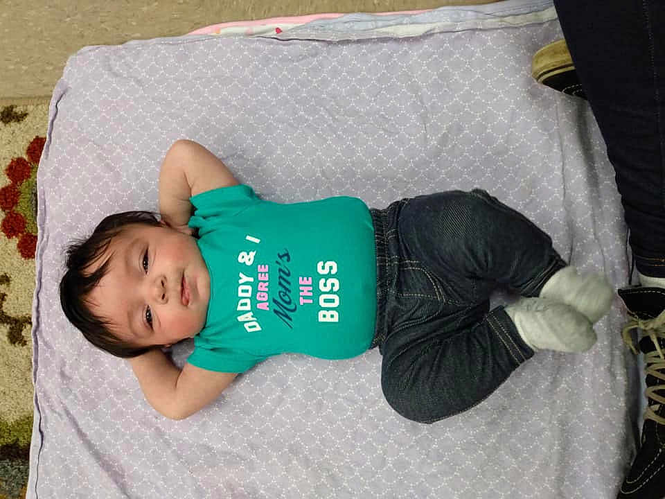 Echo is registered to the contest to win money with this photo: baby, blanket, clothing, dark_pants, face, feet, floor, footwear, happy, infant, jeans, lying_down, person, portrait, resting, shirt, socks, text_on_clothing, young_child, indoor