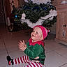 toddler, child, christmas_tree, holiday, festive, elf_costume, clapping, happy, indoor, floor, tile_floor, home, decorations, ornaments, green, red, white, striped_pants, hat, furniture
