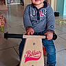 Lenny participe au concours pour gagner de l'argent avec cette photo : toddler, child, balance_bike, indoor, floor_tiles, gray_shirt, blue_jeans, black_shoes, toy, wooden_bike, smiling, person, home, playful, young_child, handlebars, floor, furniture, doorway, casual_clothing