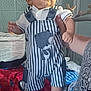 Valentin a rejoint le concours — aidez-le/la à gagner de superbes lots ! adult, baby, bear_patch, bed, blanket, blue, child, furniture, hand, holding, indoor, patterned_blanket, person, red, smiling, socks, standing, striped_overalls, tiled_wall, white_shirt
