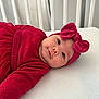 baby, infant, red_clothing, headband, bow, crib, white_bed, cute, portrait, child, smile, lying_down, soft_lighting, indoors, toddler, face, person, apparel, cozy, sleeping_area