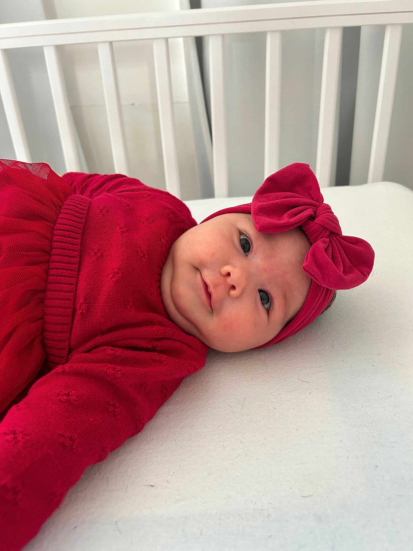 Izabella is registered to the contest to win money with this photo: baby, infant, red_clothing, headband, bow, crib, white_bed, cute, portrait, child, smile, lying_down, soft_lighting, indoors, toddler, face, person, apparel, cozy, sleeping_area