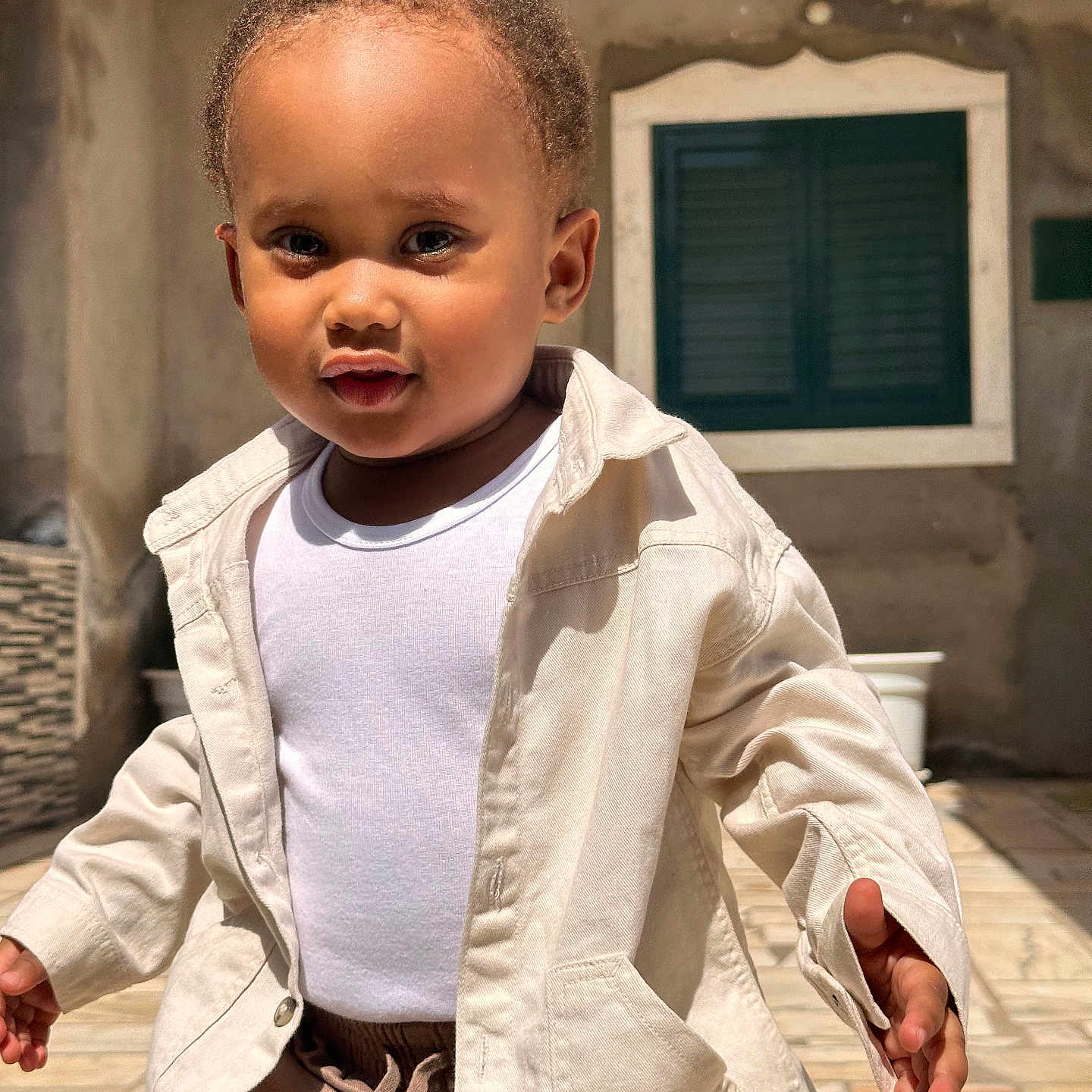 Hadriel participe au concours pour gagner de l'argent avec cette photo : casual_clothing, child, curious, curly_hair, cute, daytime, expression, face, jacket, outdoor, pavement, person, portrait, sunlight, toddler, walking, wall, white_shirt, window, young_child