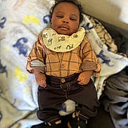 Messiah is registered to the contest to win money with this photo: baby, infant, child, face, curly_hair, bib, plaid_shirt, pants, booties, hands, blanket, patterned_blanket, sofa, indoors, portrait, cute, dapper, cozy, lying_down, smile