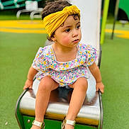Meriem participe au concours pour gagner de l'argent avec cette photo : toddler, child, playground, slide, yellow_headband, floral_dress, sandals, outdoor, green, play_equipment, summer, cute, sitting, curious, young_child, park, feet, person, daytime, playtime