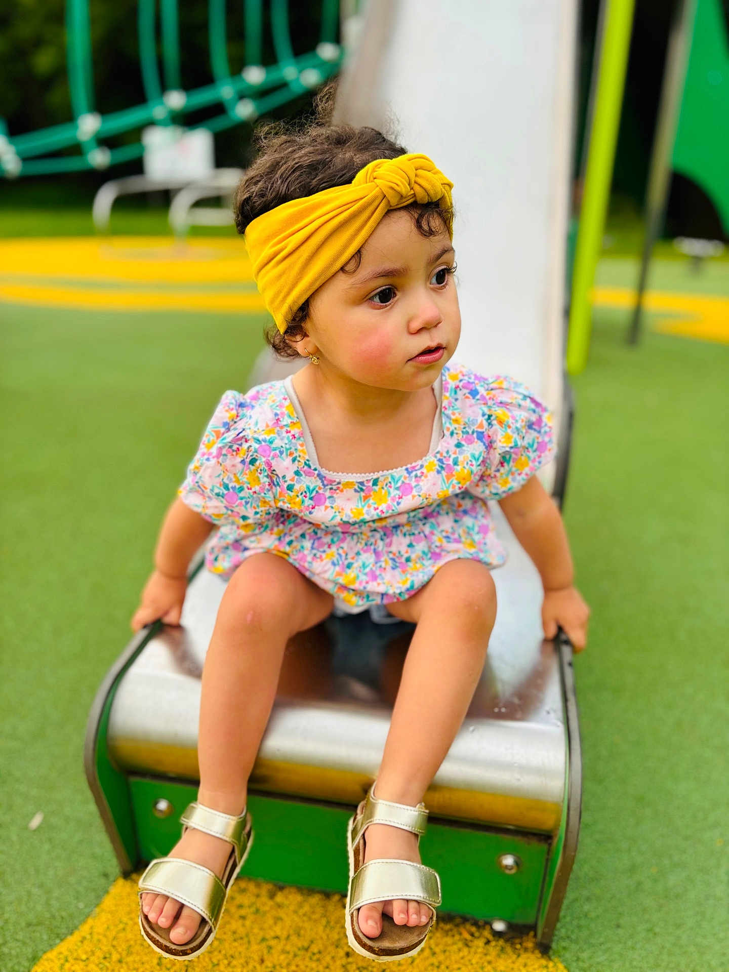 Meriem participe au concours pour gagner de l'argent avec cette photo : toddler, child, playground, slide, yellow_headband, floral_dress, sandals, outdoor, green, play_equipment, summer, cute, sitting, curious, young_child, park, feet, person, daytime, playtime