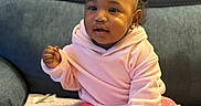 Amora is registered to the contest to win money with this photo: baby, child, pink_hoodie, floral_pants, white_shoes, sitting, blanket, couch, indoor, curly_hair, cute, smiling, face, hand, footwear, cozy, portrait, young_child, home, comfortable