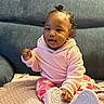 baby, child, pink_hoodie, floral_pants, white_shoes, sitting, blanket, couch, indoor, curly_hair, cute, smiling, face, hand, footwear, cozy, portrait, young_child, home, comfortable