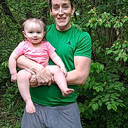Ariana is registered to the contest to win money with this photo: adult, child, toddler, person, smile, greenery, forest, outdoor, nature, holding, happy, casual_clothing, shorts, tshirt, pigtails, grass, trees, daylight, family, portrait