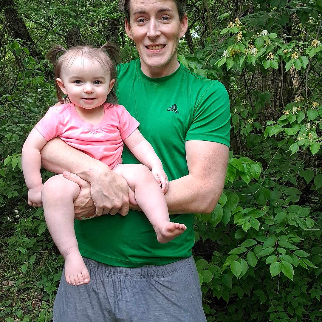 Ariana is registered to the contest to win money with this photo: adult, child, toddler, person, smile, greenery, forest, outdoor, nature, holding, happy, casual_clothing, shorts, tshirt, pigtails, grass, trees, daylight, family, portrait