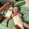baby, cat, child_hand, green_towel, bed, petting, happy, smiling, infant, animal, domestic_cat, white_clothing, relaxed, cute, indoor, person, furniture, playful, friendship, cozy