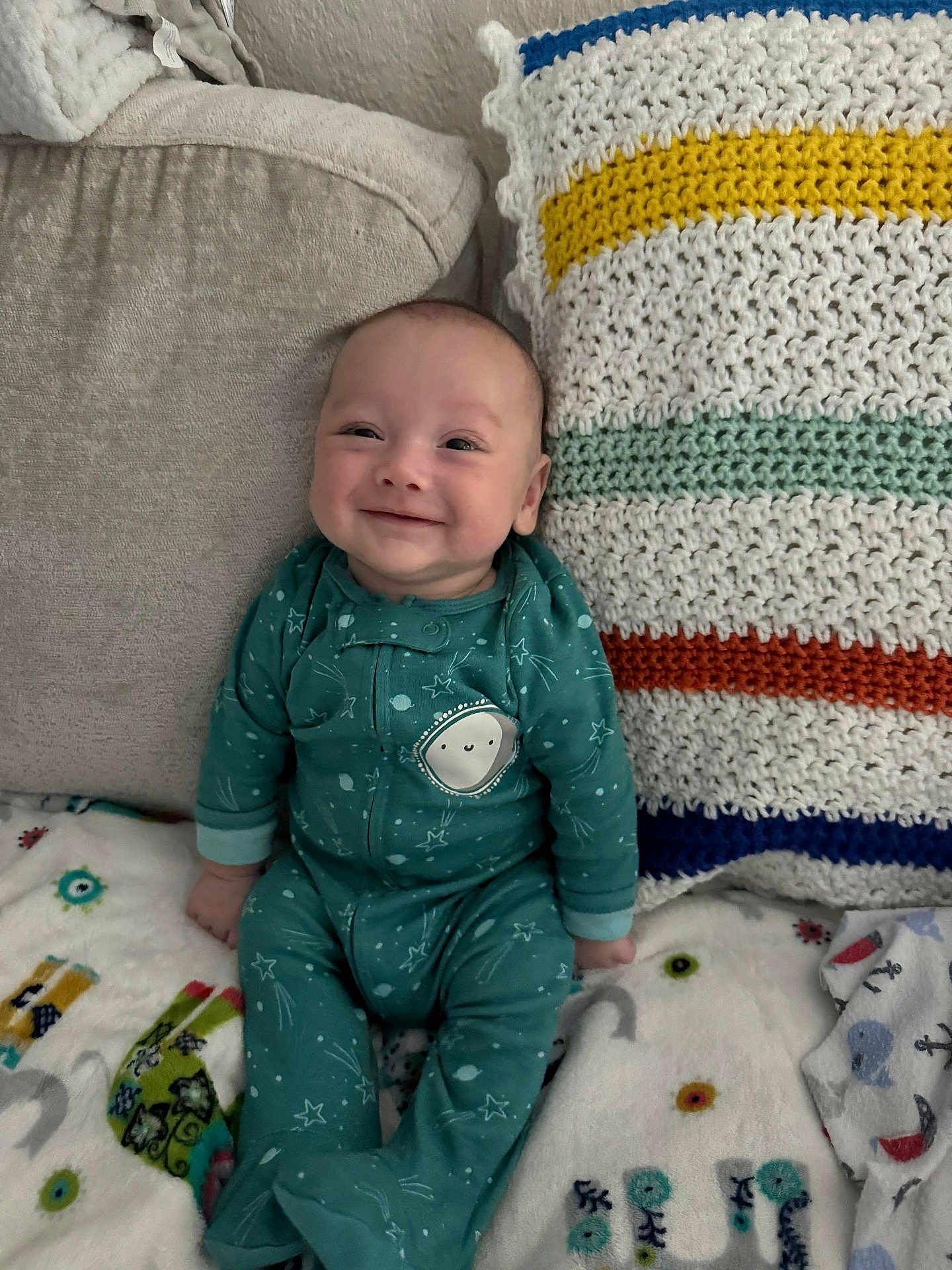 Leo is registered to the contest to win money with this photo: baby, infant, smiling, onesie, pajamas, blanket, couch, pillow, crochet_pillow, knitted_blanket, happy, portrait, cozy, seated, face, head, hands, patterned_blanket, living_room, cute