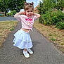 Violet is registered to the contest to win money with this photo: child, girl, pigtails, pink_shirt, white_skirt, shoes, sidewalk, trees, grass, suburban, residential_area, smiling, posing, daylight, outdoor, blue_sky, greenery, casual_clothing, happy, person
