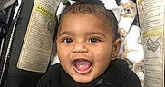 Lilly joined the competition — help win amazing prizes! baby, child, smile, black_clothing, stroller, blanket, face, happy, infant, cute, portrait, sitting, indoors, young_child, head, person, curls, bow, cozy, safety_labels